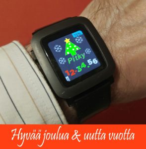 Ranteessa älykello. Sen suorakulmaisessa näytössä mustalla taustalla vihreä Pitkyn logo, jonka nimen päällä oleva muoto muutettu yksinkertaiseksi joulukuuseksi. Kuusen huipussa on tähti ja vierellä lumihiutaleita, ja alareunassa kellonaika.